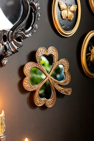 The Lucky Clover Small Gold Wall Mirror, hung on a black wall with vintage-style black and gold wall art. 