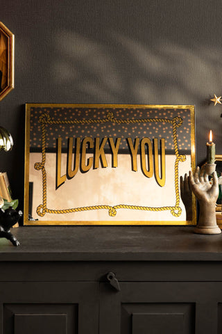 The Lucky You Mirror shown on a black sideboard with gold accessories. 