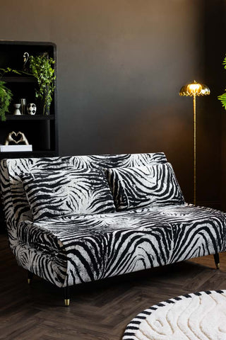 The Mabel Large 2 Seater Sofa Bed in Zebra Print Velvet shown against dark walls with a shelf in the background and a floor lamp in the corner. 