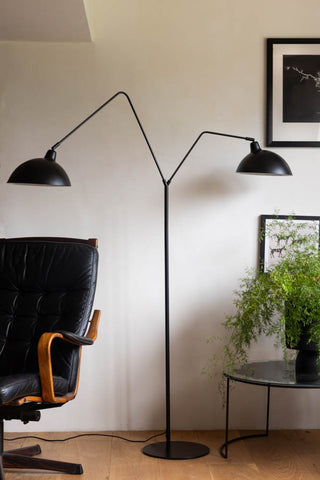 Lifestyle image of the Matt Black Double Floor Lamp displayed on a wooden floor in front of a white wall, surrounded by various pieces of furniture, home accessories and plants. 