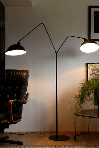 Lifestyle image of the Matt Black Double Floor Lamp displayed illuminated on a wooden floor in front of a white wall, surrounded by various pieces of furniture, home accessories and plants.