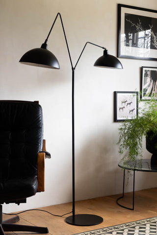 Lifestyle image of the Matt Black Double Floor Lamp displayed on a wooden floor in front of a white wall, surrounded by various pieces of furniture, home accessories and plants.