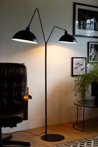 Lifestyle image of the Matt Black Double Floor Lamp displayed illuminated on a wooden floor in front of a white wall, surrounded by various pieces of furniture, home accessories and plants.