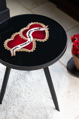 The Milagro Heart Round Side Table seen from above, styled on a rug with a potted plant in the background.