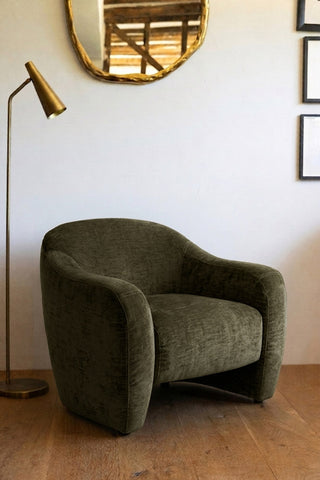 The Modern Curved Armchair in Olive Green styled next to a lamp, in front of a wall with a mirror and some framed artwork on.