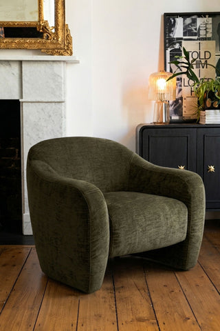 The Modern Curved Armchair in Olive Green styled in a living room with various accessories in the background.