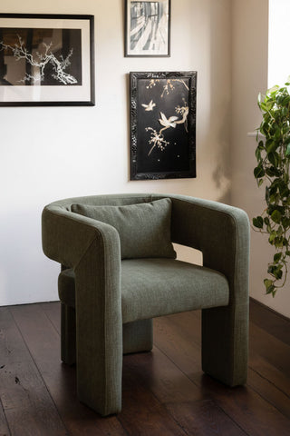 The Modern Green Accent Chair with Bolster Cushion styled in the corner of a room with some framed artwork and a plant.