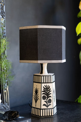 The Monochrome Folklore Floral Table Lamp shown with the light off against a black wall with plants in the background. 