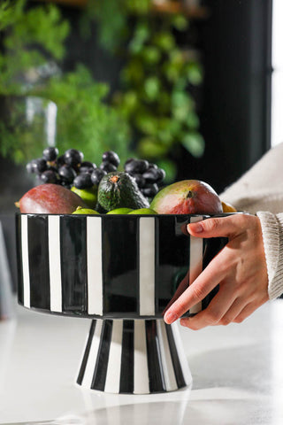 Lifestyle image of the Black & White Stripe Bowl with fruit inside, being placed onto a table by someone's hands. 