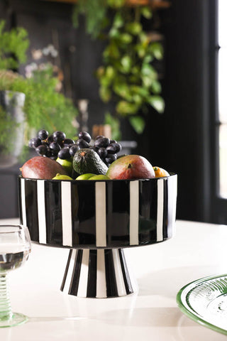 Lifestyle image of the Black & White Stripe Bowl displayed on a white kitchen table with fruit inside, styled with other tableware. 