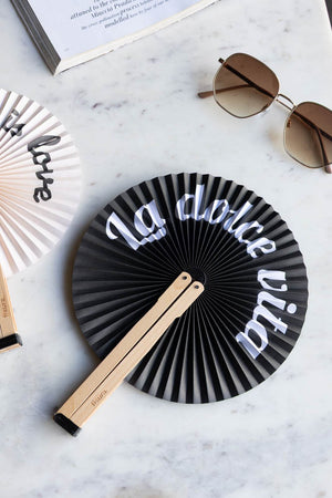 The Monochrome La Dolce Vita Hand Held Fan on a marble surface with another fan, some sunglasses and a book.