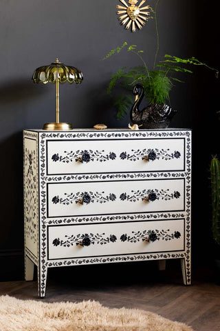 The Monochrome Rose Noir Chest Of Drawers in front of a dark wall, styled with various plants and accessories.