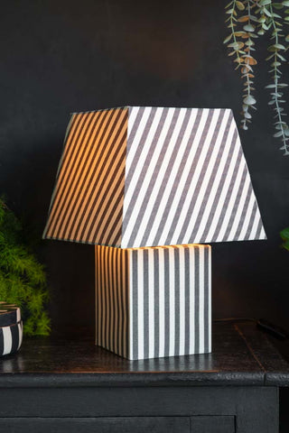 The Monochrome Stripe Table Lamp switched on and displayed on a black sideboard with some plants.