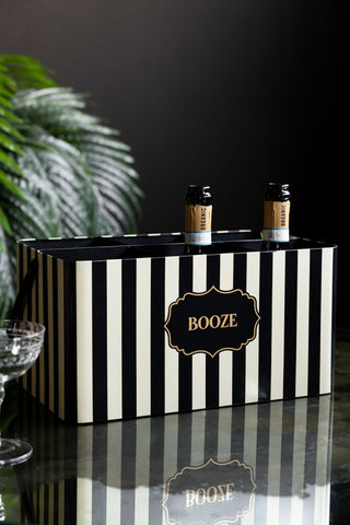 The Monochrome Stripe "Booze" Wine Bottle Carrier with some bottles inside, styled on a dark surface with a champagne glass and some greenery.