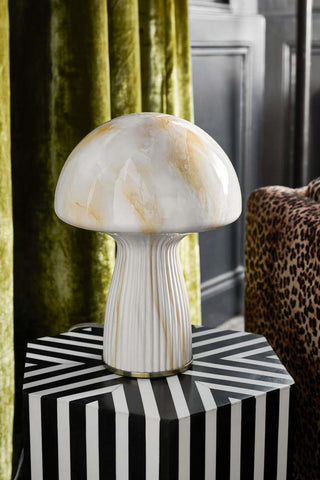 Lifestyle image of the White Porcini Table Lamp displayed on a black and white side table, next to a leopard print chair and green velvet curtains.
