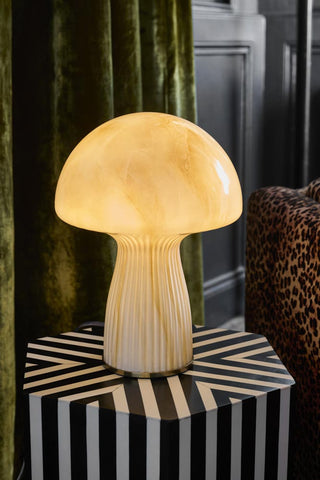 Lifestyle image of the White Porcini Table Lamp displayed illuminated on a black and white side table, next to a leopard print chair and green velvet curtains.