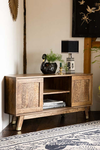 The Natural Leopard Print Corner TV Unit shown in the corner of a neutral room with a black swan plant pot and monochrome folk table lamp styled on top of the surface. 