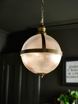Lifestyle image of the Boulevard Pendant Light illuminated and displayed in a black room, with a plant and art print in the background. 