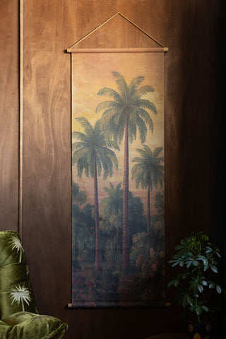 The Palm Tree Canvas Wall Hanging on a dark wooden wall, above a plant and a green velvet chair.