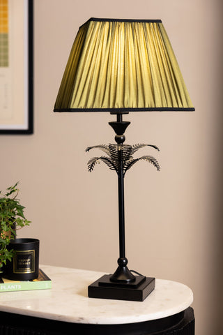 The Palm Tree Table Lamp - Green & Black, switched on and displayed ona marble surface with some other accessories.