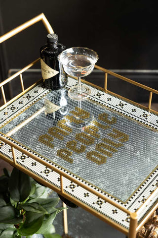 The Party People Only Gold Drinks Trolley, displaying the slogan on the top shelf with a champagne glass and bottle for decoration, and a plant on the bottom shelf on a dark background. 