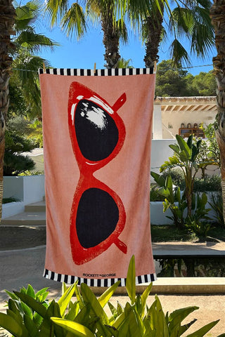The Pink Glasses Beach Towel displayed hanging in the sunshine surrounded by trees and plants.
