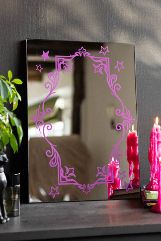 The Pink Ornate Print Frame Mirror shown against a black wall with pink candles in the background. 