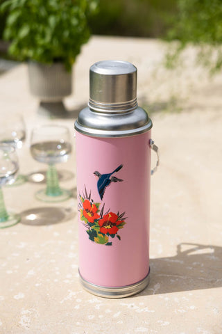 Lifestyle image of the Pink Painted Bird Decorative Flask on an outdoor table with some water glasses and plants in the background.