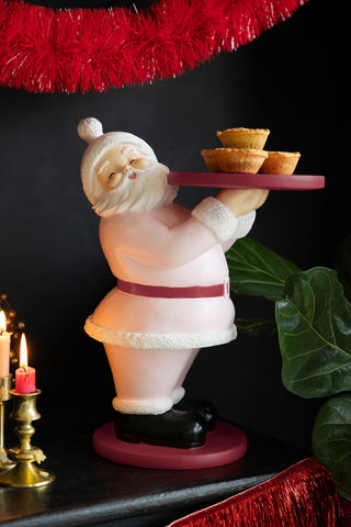 The Pink Santa Butler Christmas Decoration holding some mince pies, displayed on a black surface with various decorations.