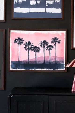 Framed artwork of palm trees on a pink and blue background against a dark wall.