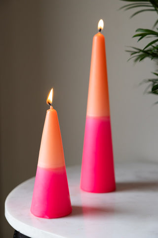 The Small and Large Pink & Orange Cone Shaped Candles displayed lit together on a white table with the leaves of a plant in the background.