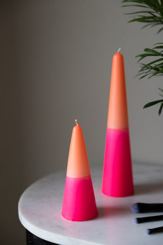 The Small and Large Pink & Orange Cone Shaped Candles displayed together on a white marble table next to some makeup brushes, with a plant in the background.