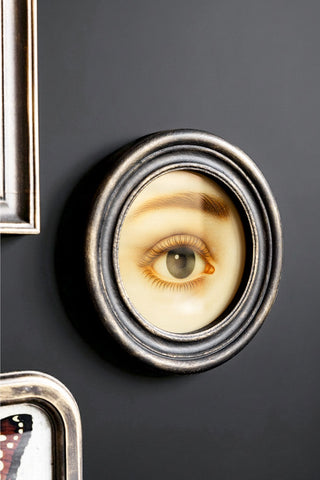The Vintage-Style Pretty Eye Framed Wall Art seen from an angle, displayed on a black wall with two other artworks.