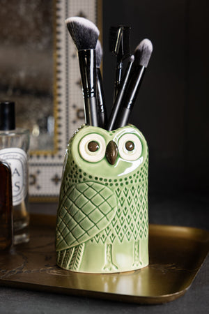 The Quirky Green Owl Bud Vase styled with makeup brushes inside, displayed on a trinket tray with some perfume.