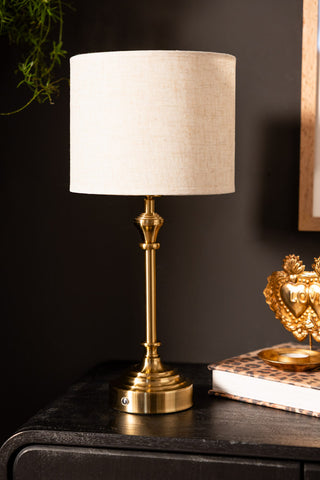 The Rechargeable Antique Bronze Table Lamp with Neutral Shade displayed on a black sideboard with a leopard print book and a tealight holder.