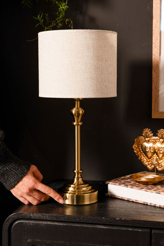 Person turning on a brass table lamp with a white shade on a dark surface.