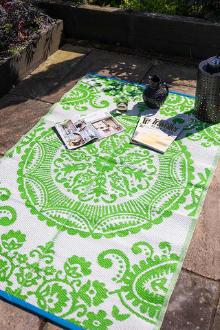 The Recycled Vintage Design Outdoor Rug in Green styled on a patio in the sunshine with a solar lantern, jug, glass and some magazines.