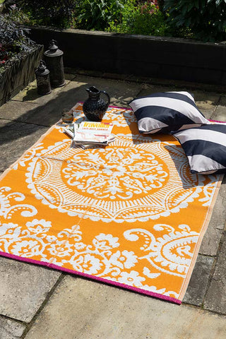 The Recycled Vintage Design Outdoor Rug in Orange on a patio in the sun, styled with monochrome striped cushions, magazines, a jug, a glass and some lanterns.