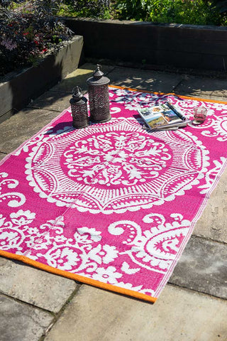 The Recycled Vintage Design Outdoor Rug in Pink on a patio in the sunshine, styled with lanterns, magazines and a glass.
