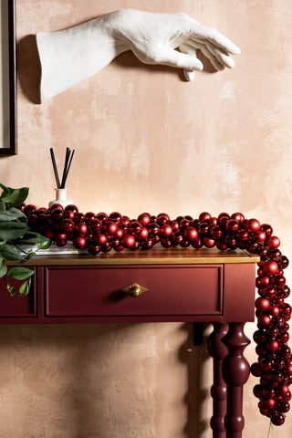 The Red Bauble Cluster Christmas Garland draped across a red console table, styled with a diffuser, greenery and a hand wall sculpture.