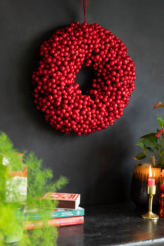 The Red Berry Christmas Wreath styled on a black wall above various accessories and a lit candle.