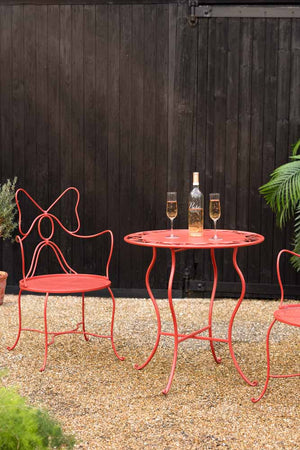The Red Bow Folding Garden Table & Chairs Set with a bottle of wine and some glasses on, styled on an outdoor patio with some plants.