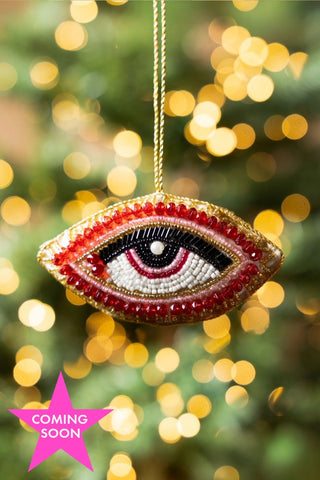 The Red Eye Beaded Hanging Christmas Tree Decoration hanging in front of blurred fairy lights. Image includes a pink star icon with the words 'coming soon'.
