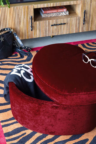 The Red Velvet Heart Footstool With Storage with the lid off showing the storage inside, displayed on a patterned rug with a tv unit in the background.