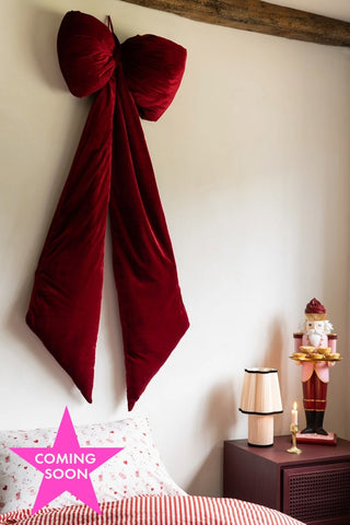 The Red Velvet Oversized Bow Decoration on the wall above a bed shown with a pink star icon that reads 'coming soon'.