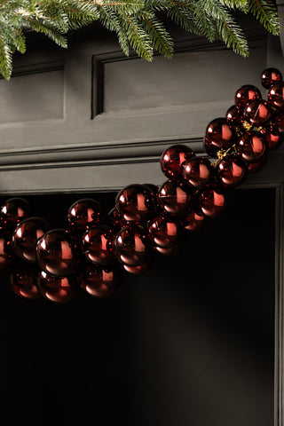 The Red Antique-Effect Bauble Christmas Garland styled on the edge of a black fireplace with some greenery.