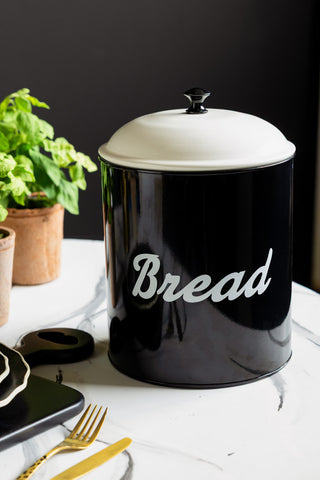 The Retro Monochrome Enamel Bread Bin shown in a black kitchen on a marble table. The bread board has a black base with "bread" written across it and a white lid with a black handle. 