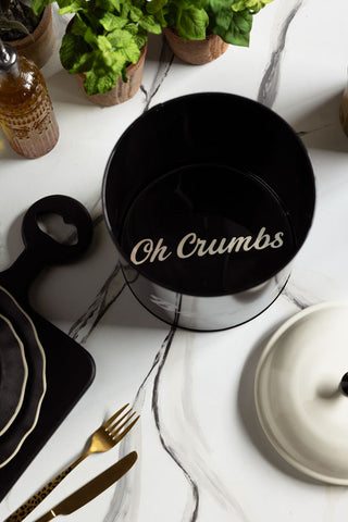 The inside of the Retro Monochrome Enamel Bread Bin, showing the words "oh crumbs" hidden inside. Bread board is shown on a marble worktop with plants and a bread board in the background. 