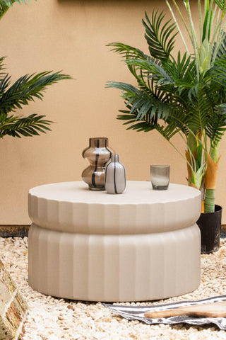 The Ribbed Edge Round Coffee Table In Ivory styled outdoors with some vases and a tumbler on, surrounded by plants.