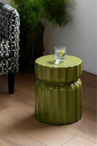 The Ribbed Edge Round Side Table in Green Glaze displayed with a glass tumbler on, with a chair and a plant in the background.